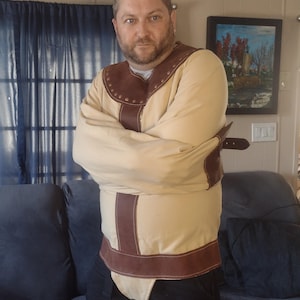 Könnte beinhalten: Eine beige und braune Zwangsjacke mit Lederdetails. Das Kleidungsstück hat lange Ärmel und einen braunen Lederkragen. Die Vorderseite der Jacke hat braune Lederriemen und einen Schnallenverschluss. Die Person trägt eine schwarze Hose.