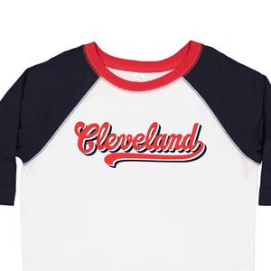 May include: White baseball tee with navy blue sleeves and a red collar. The front of the shirt has a red and blue graphic that says "Cleveland" in a retro font.
