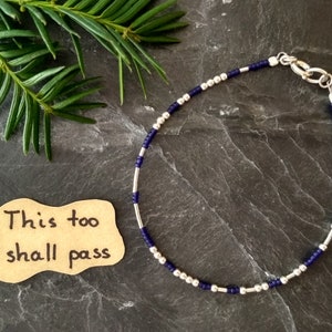Könnte beinhalten: Ein filigranes Armband mit abwechselnd silbernen und dunkelblauen Perlen. Das Armband ist mit einem silbernen Verschluss versehen. Eine kleine, handgeschriebene Notiz mit dem Text "This too shall pass" ist ebenfalls sichtbar.