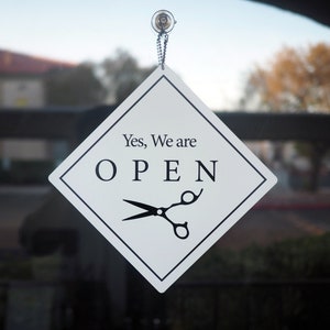 May include: A white diamond-shaped sign with black trim hangs on a glass door. The sign reads "Yes, We are OPEN" with a pair of black scissors underneath the text.