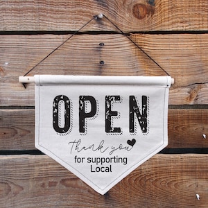 May include: A white canvas sign with black text that reads "OPEN" and "Thank you for supporting Local" with a small black heart. The sign is hanging from a wooden wall.