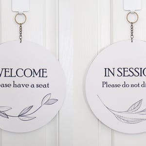 Può includere: Due cartelli rotondi bianchi con testo nero e un semplice disegno a linea nera di un ramo con foglie. Il cartello a sinistra dice "Benvenuti, Prego accomodatevi". Il cartello a destra dice "In sessione, Si prega di non disturbare".