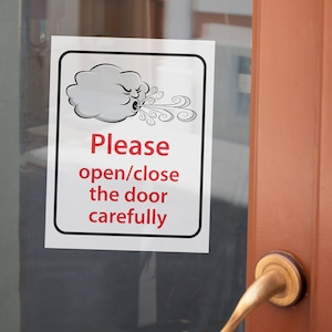 May include: A white sign with black trim and red text that reads "Please open/close the door carefully". A cartoon image of a cloud blowing wind is above the text.