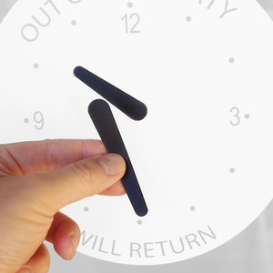 Puede incluir: Una esfera de reloj blanca con agujas negras ajustadas a la 1:55. La esfera del reloj tiene las palabras "OUT TO" en la parte superior y "WILL RETURN" en la parte inferior.