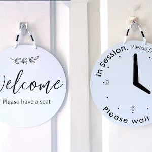 May include: Two round white signs with black text and graphics. The first sign says "Welcome" with a small branch of leaves and "Please have a seat". The second sign says "In session. Please Do Not Disturb. Please wait until" with a clock face and a black hand pointing to the 4 o'clock position.