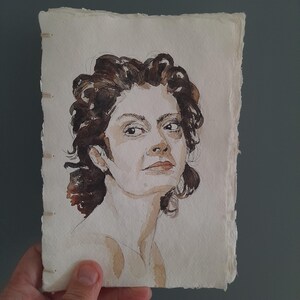 May include: A watercolor portrait of a person with dark, curly hair painted on a rectangular piece of off-white, textured paper with deckled edges. The artwork features visible brushstrokes and ink outlines, bound on the left side with light-colored thread.