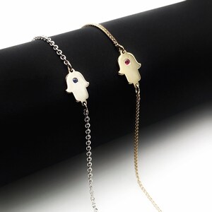 May include: Two delicate hamsa hand bracelets with a chain link design. One bracelet has a blue gemstone in the center of the hamsa hand, and the other has a red gemstone.