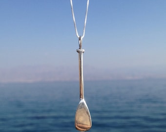 May include: A silver paddle pendant necklace hanging against a blue sky and ocean background.