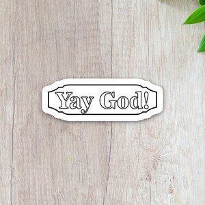 May include: A white sticker with black text that reads "Yay God!" The sticker is shaped like a banner with a decorative border.