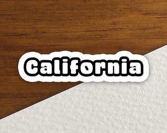 May include: A white sticker with the word "California" in a bold, black font. The sticker has a black border and is set against a wooden surface and a white paper background.
