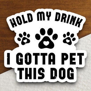 Peut inclure: Autocollant blanc avec du texte noir qui dit "Hold my drink I gotta pet this dog". Trois empreintes de pattes noires avec un cœur rouge au centre sont au-dessus du texte.