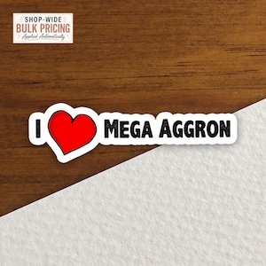 May include: A rectangular sticker featuring the text I, a red heart symbol, and the text MEGA AGGRON in black capital letters. The sticker has a white border and is positioned across a surface that is half dark wood grain and half white texture.
