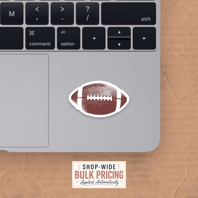May include: A brown and white American football sticker with a textured, distressed appearance. The sticker is placed on the surface of a silver laptop keyboard. Below the laptop, a white rectangular label displays the text SHOP-WIDE BULK PRICING.