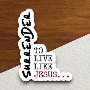 May include: Black and white sticker with the text "Surrender to live like Jesus..." in a vertical orientation. The word "Jesus" is in red.