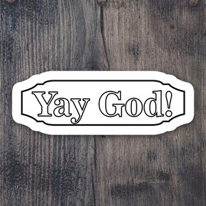 May include: White sticker with black outline and the text "Yay God!" on a dark brown wooden background.