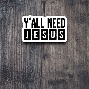Peut inclure: Un autocollant blanc avec du texte noir sur un fond en bois. L'autocollant dit "Y'all need Jesus" avec le mot "Jesus" dans des carr&eacute;s individuels.