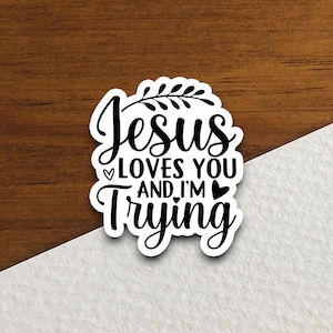 Op de afbeelding: Een witte sticker met zwarte tekst: "Jesus loves you and I'm trying." De sticker heeft een zwart bladontwerp en hartafbeeldingen. Op een houten achtergrond.