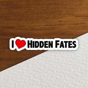 May include: A white sticker with the text "I ❤️ Hidden Fates." The heart is red, and the text is in a bold black font. The sticker is on a wooden surface with a white paper background.