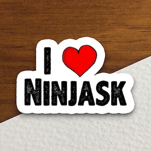 Pode incluir: Um autocolante branco com as palavras "I" e "NINJASK" em preto, com um coração vermelho entre elas. O autocolante tem uma borda preta e está numa superfície de madeira.