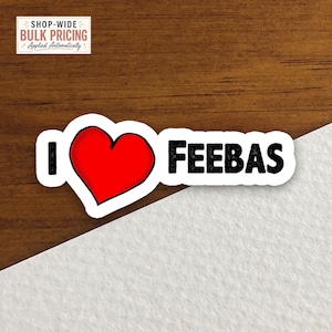 May include: A die-cut sticker featuring the text I, a large red heart symbol, and the text FEEBAS in bold black capital letters. The sticker has a white border and is displayed against a split background of dark wood grain and white texture.