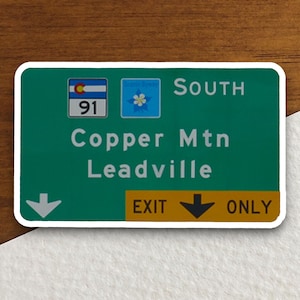 Puede incluir: Una señal de carretera verde con borde blanco, que indica las direcciones a Copper Mtn y Leadville, Colorado. La señal presenta una flecha blanca apuntando hacia abajo, el texto "EXIT ONLY" y el número "91" en un círculo azul. La señal también incluye un pequeño cuadrado azul con una flor blanca, que indica una carretera panorámica.