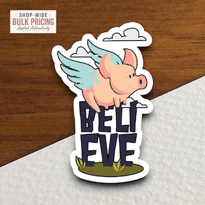 May include: A die-cut sticker featuring a light pink pig with light blue wings flying above the dark purple text BELIEVE. The design includes white clouds in the background and a small patch of green grass at the base of the bold lettering.