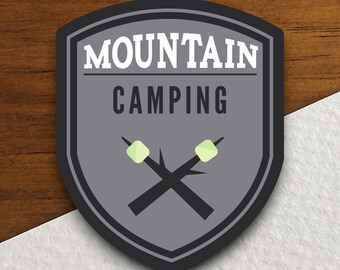 Könnte beinhalten: Ein grauer, schildförmiger Patch mit dem Text "MOUNTAIN CAMPING" in Weiß. Zwei gekreuzte Stöcke mit weißen Marshmallows an den Enden befinden sich in der Mitte des Patches.
