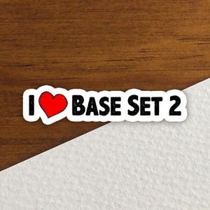 May include: A sticker with the phrase "I ❤️ BASE SET 2" in black text with a red heart. The sticker has a white border and is set against a brown wooden surface and a white surface.