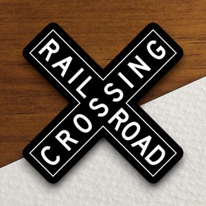 Peut inclure: Panneau de passage à niveau noir et blanc en forme de croix. Les mots "RAILROAD CROSSING" sont imprimés en blanc, avec un bord noir. Le panneau est posé sur un fond en bois.