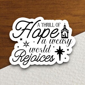 May include: A white sticker with black text and a black silhouette of a nativity scene. The text reads "A thrill of hope a weary world rejoices".