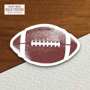 May include: A brown American football sticker with a white lace pattern and white stripes on each end. The sticker features a textured, grainy surface design and is die-cut with a white border, showcasing a classic sports ball graphic element.