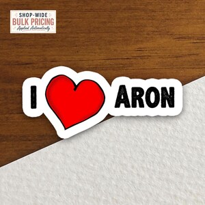 May include: A die-cut sticker featuring the text I, a bright red heart symbol, and the name ARON in bold black capital letters. The sticker has a white border and is displayed against a split background of dark wood grain and white paper.