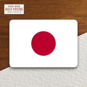 May include: A rectangular sticker featuring the national flag of Japan, which consists of a white background with a solid red circle centered in the middle. The sticker has rounded corners and is displayed against a contrasting wood and white surface.
