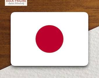 May include: A rectangular sticker featuring the national flag of Japan, which consists of a white background with a solid red circle centered in the middle. The sticker has rounded corners and is displayed against a contrasting wood and white surface.