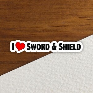 May include: A white sticker with the text "I ❤️ SWORD & SHIELD" in black, with a red heart replacing the word "love". The sticker is on a wooden surface with a white angled background.