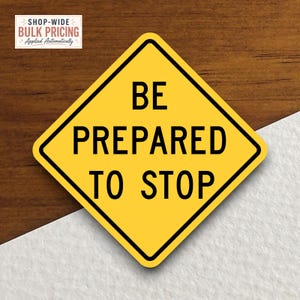 May include: A diamond-shaped yellow road sign with a black border and the black text BE PREPARED TO STOP. The sign features a standard traffic warning design with bold sans-serif lettering centered on the yellow background, mimicking a highway sign.