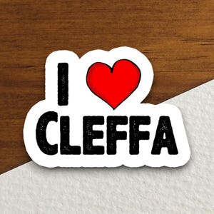 May include: A white sticker with the words "I CLEFFA" in black, bold letters. The heart is red. The sticker is on a wooden surface.