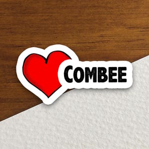 May include: A die-cut sticker featuring a vibrant red heart shape positioned to the left of the word COMBEE, which is printed in bold, black, sans-serif capital letters. The entire design has a clean white border surrounding the graphic elements.