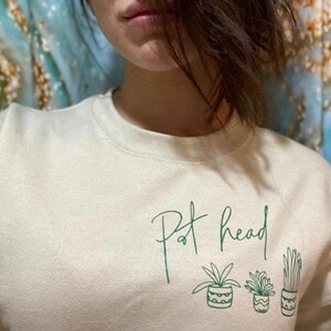 Puede incluir: Una sudadera beige claro con el texto "Pot Head" en escritura verde. Debajo del texto hay tres dibujos de plantas en maceta en línea verde.