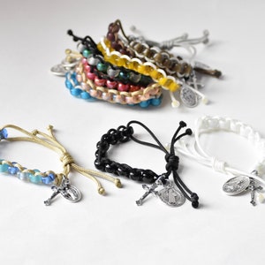 May include: A collection of adjustable cord bracelets featuring colorful beads and silver-toned metal charms, including crucifixes and religious medallions. The bracelets are arranged in a pile and individually displayed in blue, black, and white.