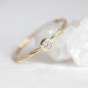 Puede incluir: Un delicado anillo de oro con un solo diamante redondo y transparente engastado en engaste de bisel. La banda está retorcida y tiene un acabado pulido.