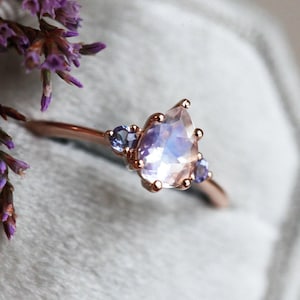 May include: A pear-shaped moonstone ring set in rose gold with two smaller blue gemstones on either side.