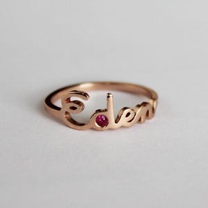 Peut inclure: Une bague en or rose avec le nom "Eden" écrit en lettres cursives. Une petite pierre précieuse rubis est sertie au centre de la bague.