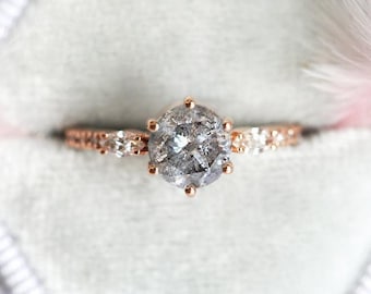 Puede incluir: Un anillo de compromiso de oro rosa con un gran diamante gris redondo. El diamante tiene un aspecto único, crudo y natural. La banda está engastada con diamantes más pequeños.