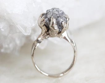 May include: A silver ring with a large, raw, grey diamond set in a claw setting.