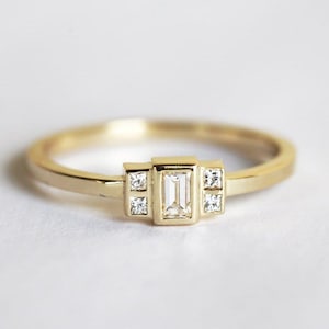 Puede incluir: Un anillo de banda de oro con un diamante de forma rectangular en el centro y dos diamantes cuadrados más pequeños a cada lado.