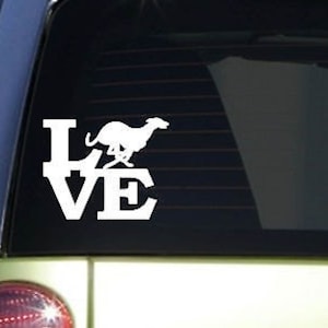 May include: White vinyl decal with the word "LOVE" and a silhouette of a running Greyhound dog. The decal is on a car window.