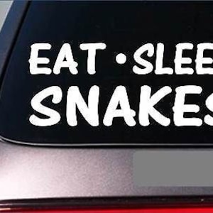 May include: White vinyl decal with the text "Eat - Sleep Snakes" on a black car window.