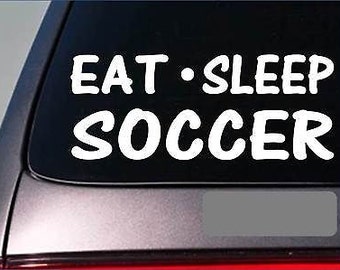 May include: White vinyl decal with the text "Eat - Sleep Soccer" on a black background. The decal is applied to the rear window of a car.