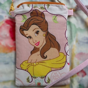 Puede incluir: Una funda para teléfono de tela rosa y blanca con una imagen de Bella de La Bella y la Bestia. La funda tiene una correa rosa.
